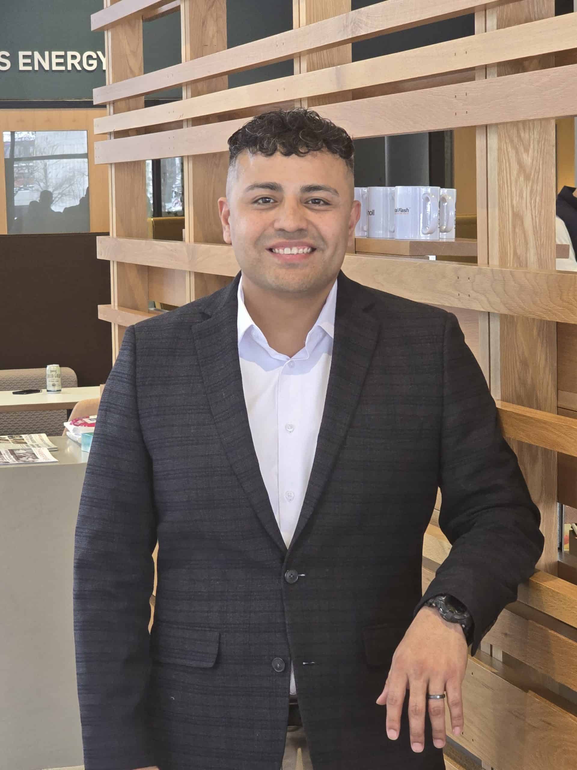Contact 1 Johnny, a professional sales representative, standing in an office setting wearing a dark checked blazer and white shirt, smiling confidently.